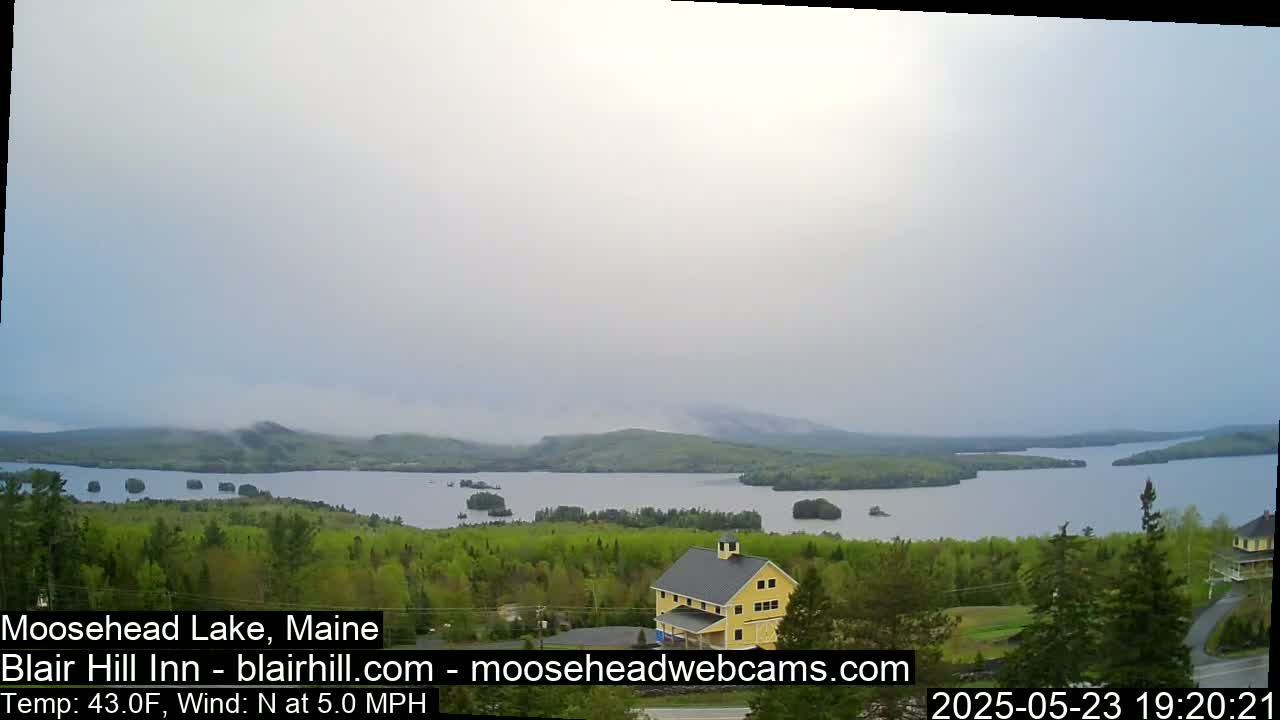 A hazy view of a lake and forested hills, with a yellow building in the foreground, under a mostly cloudy sky.