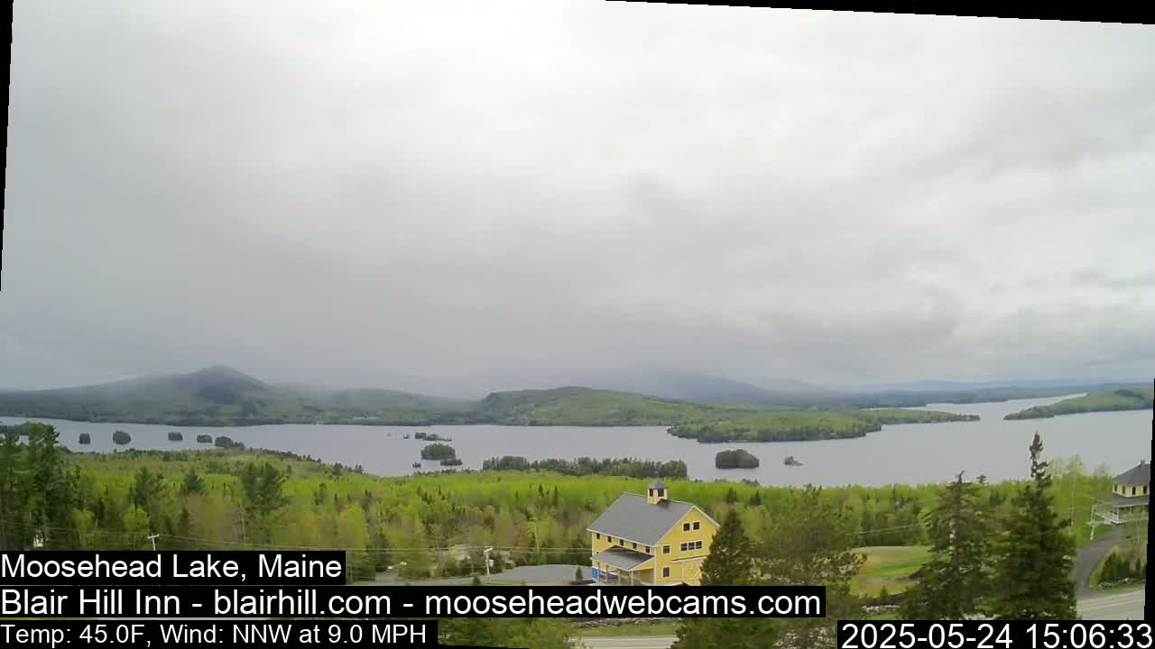 A yellow building sits on a hill overlooking a lake and forested mountains under an overcast sky.