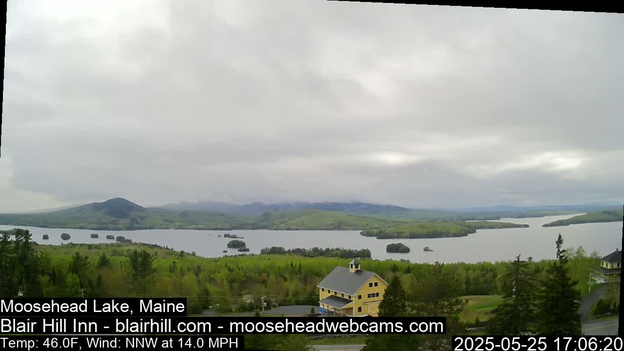 A yellow building sits on a hill overlooking a large lake under an overcast sky with a 14 mph northwest wind and a temperature of 46°F.