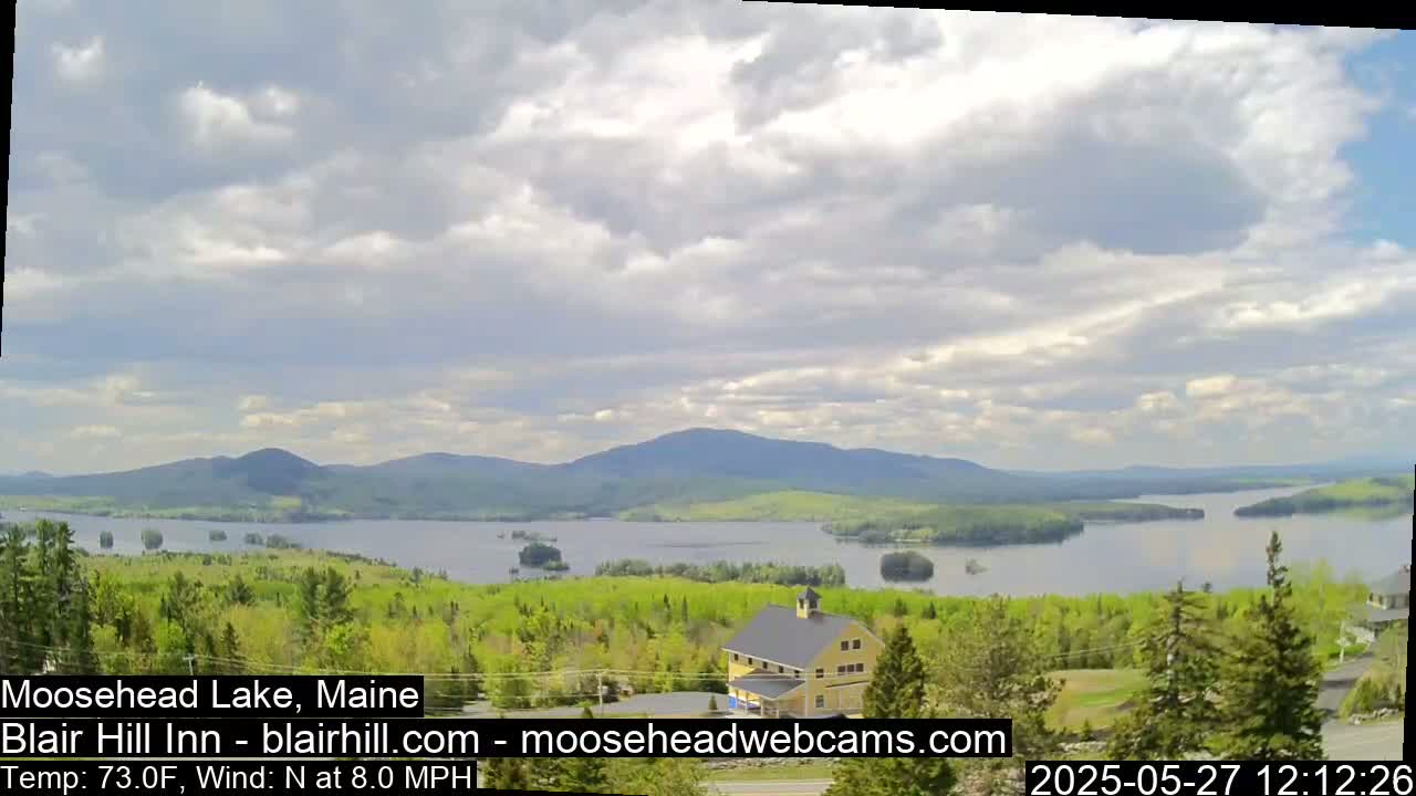 A partly cloudy day reveals a view of a lake surrounded by green trees and distant mountains, with a building visible in the foreground.