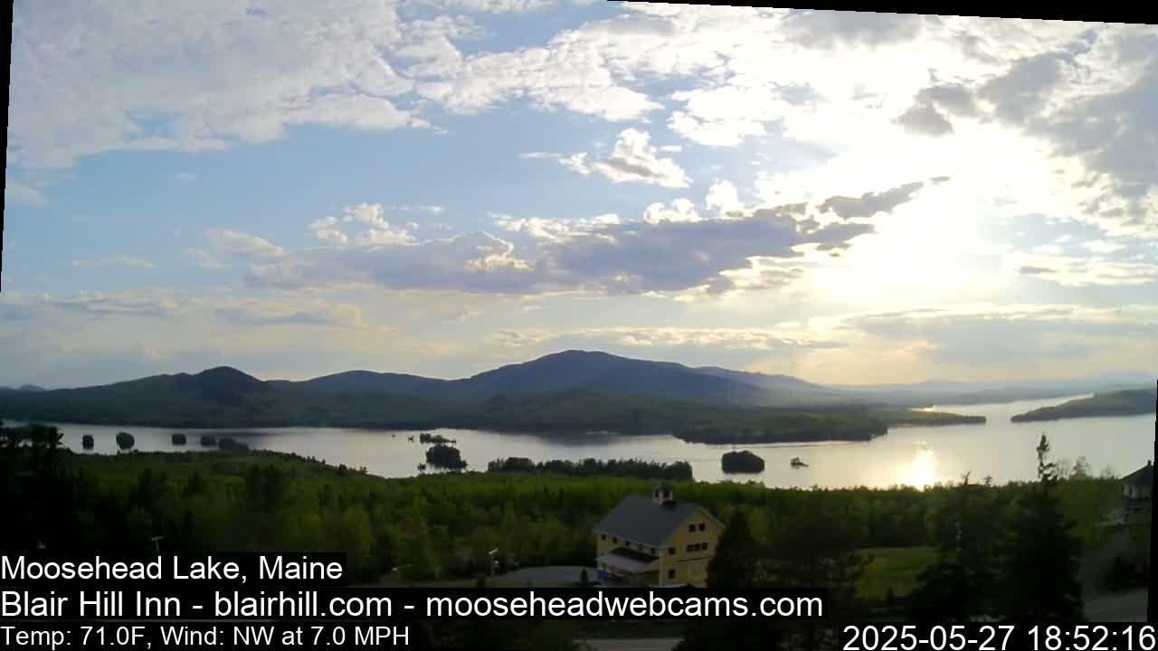 A partly cloudy sunset over a large lake nestled among rolling green hills and a single building.