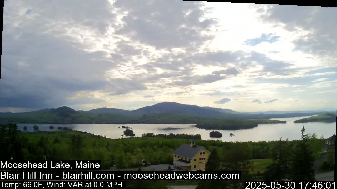 A partly cloudy sky overlooks a calm lake surrounded by green hills and a yellow building.