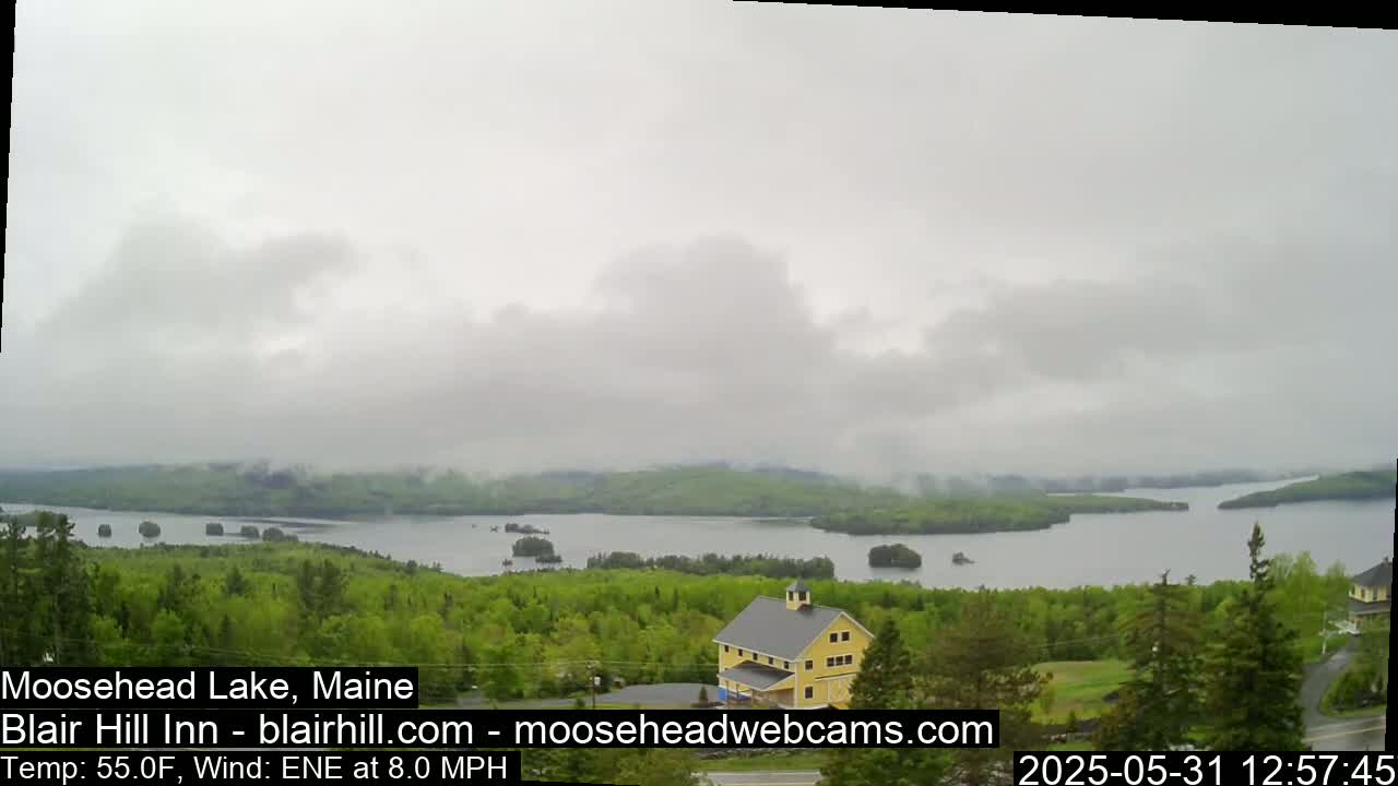 A yellow building sits atop a lush green hill overlooking a large lake under a mostly cloudy sky.