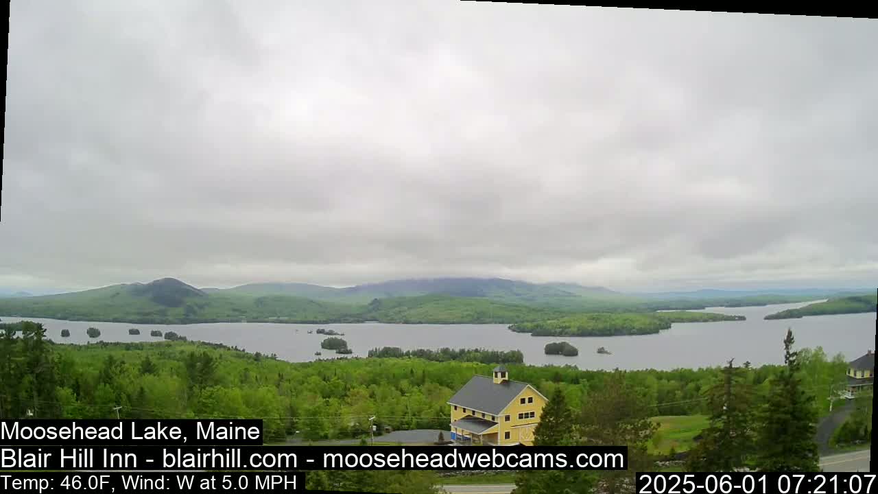 A yellow building sits on a hill overlooking a large lake and forested mountains under an overcast sky.