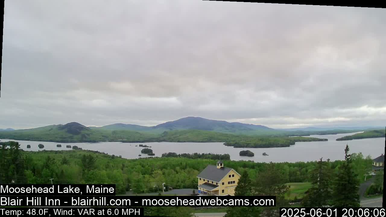 A large lake surrounded by green trees and mountains is viewed from a vantage point above, under an overcast sky.