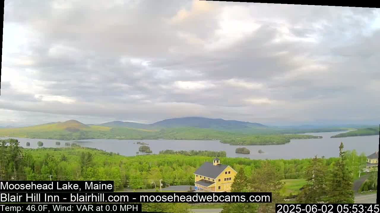 A large lake surrounded by green trees and mountains is visible under a mostly cloudy sky, with a yellow building situated in the foreground.