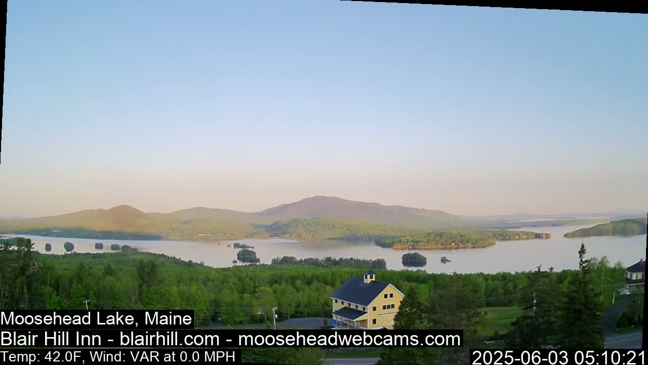 A calm, light-blue sky overlooks a tranquil lake surrounded by lush green trees and distant mountains under a clear sky.