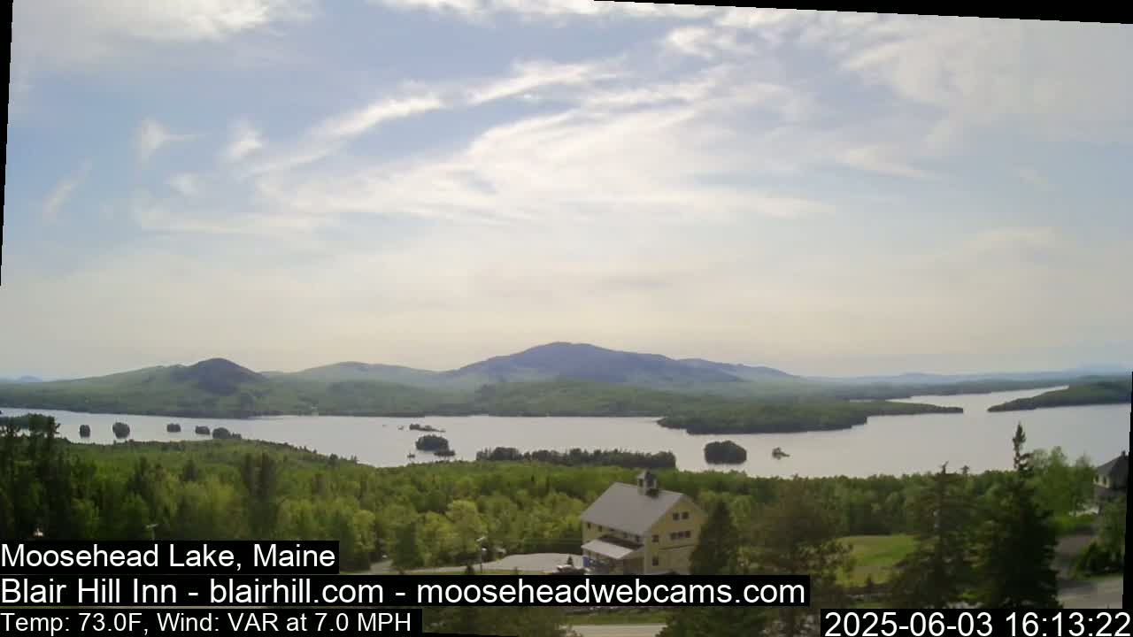 A partly cloudy day reveals a large lake nestled among green hills and a building overlooking the tranquil scene.