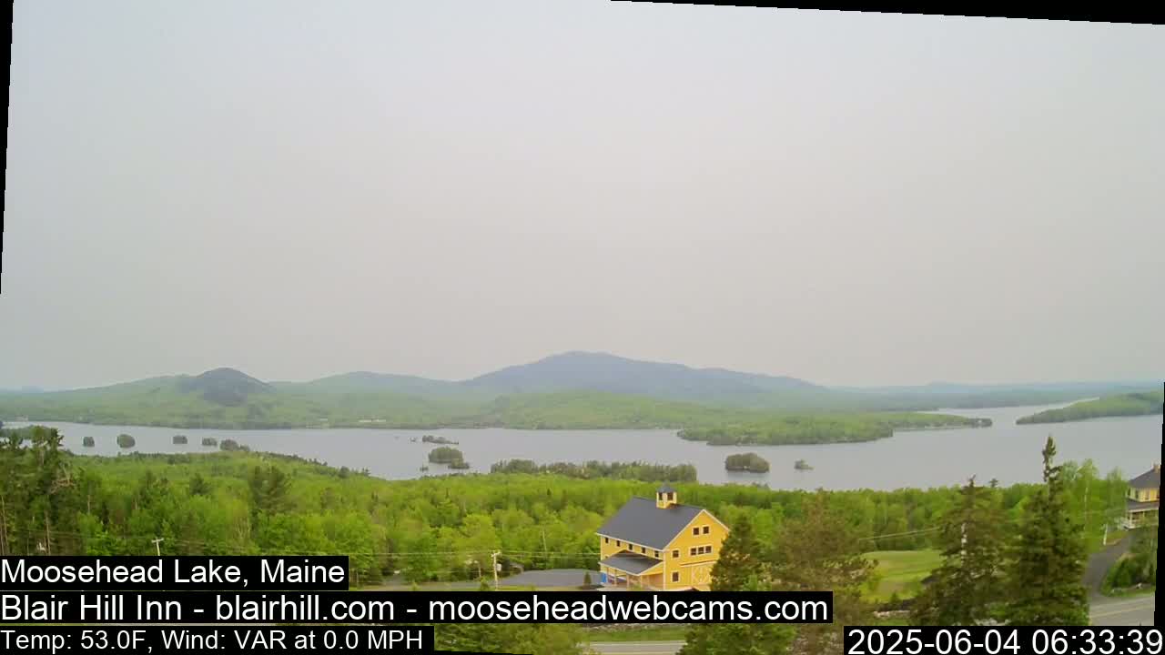 A yellow building sits on a hill overlooking a calm lake with forested hills and islands in the distance under an overcast sky.