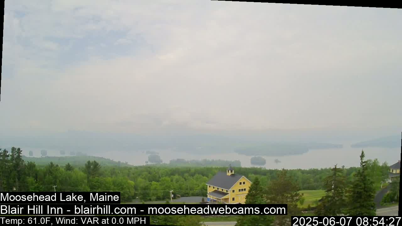 A hazy view of Moosehead Lake in Maine shows a yellow barn and green trees on a hill overlooking the calm water.