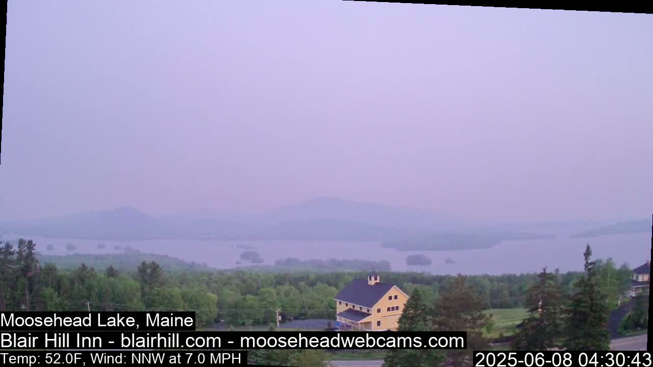 A hazy view of a lake and distant mountains is seen from a vantage point above a yellow building and green trees, under a light purple sky.