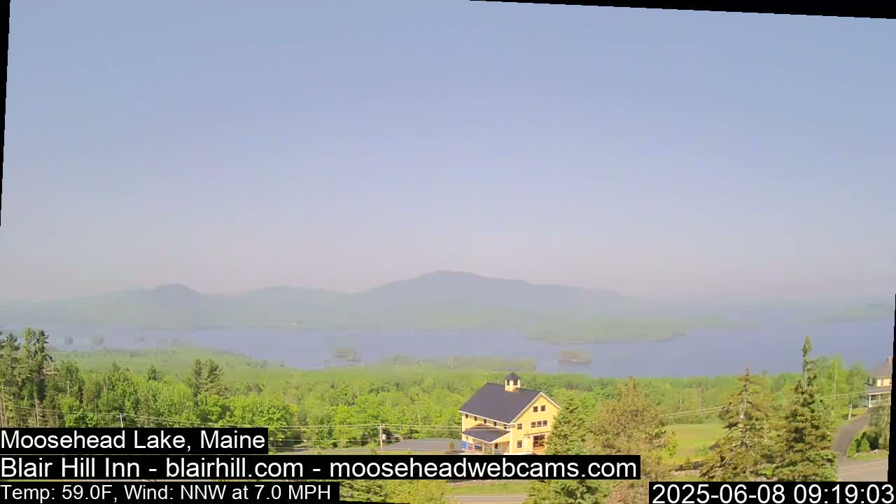 A hazy, sunny view of a lake and distant mountains from a vantage point overlooking a yellow building and lush green trees, with a light wind blowing.