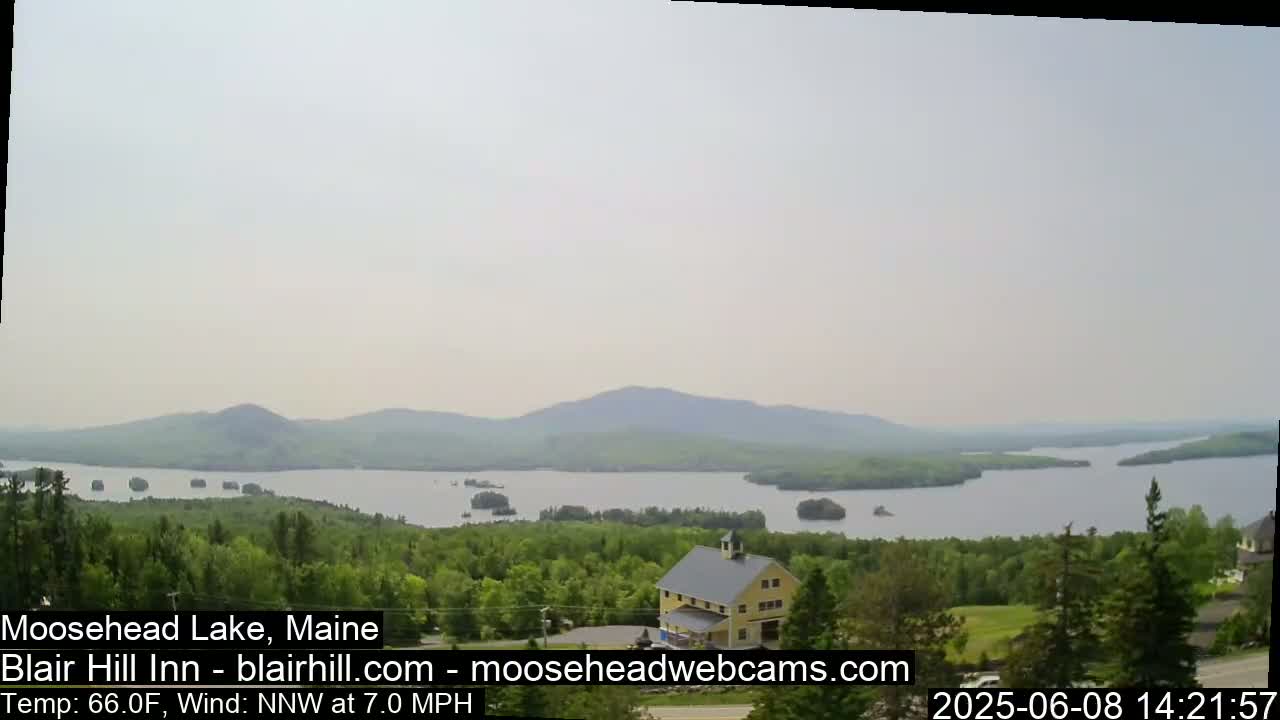 A hazy view of a large lake surrounded by green trees and mountains, with a yellow barn visible in the foreground.