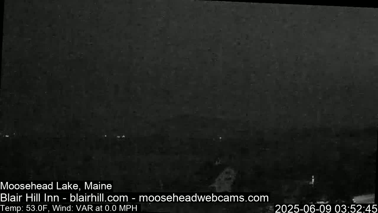 A nighttime, low-light view of a dark landscape with faint lights in the distance and a calm wind.