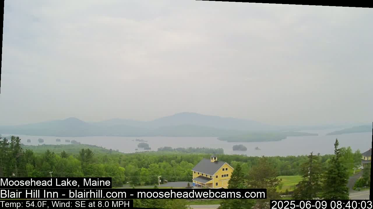 A hazy view of Moosehead Lake in Maine, showing a large body of water, distant mountains, and a yellow building nestled among green trees, with a southeast wind at 8 mph and a temperature of 54°F.