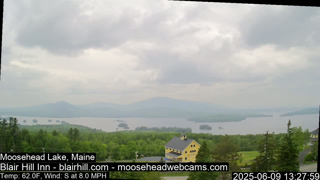 A yellow building sits on a hill overlooking a large lake and distant mountains under a cloudy sky.