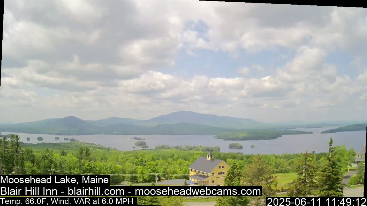 A partly cloudy day reveals a view of a lake surrounded by green trees and distant mountains, with a yellow building visible in the foreground.