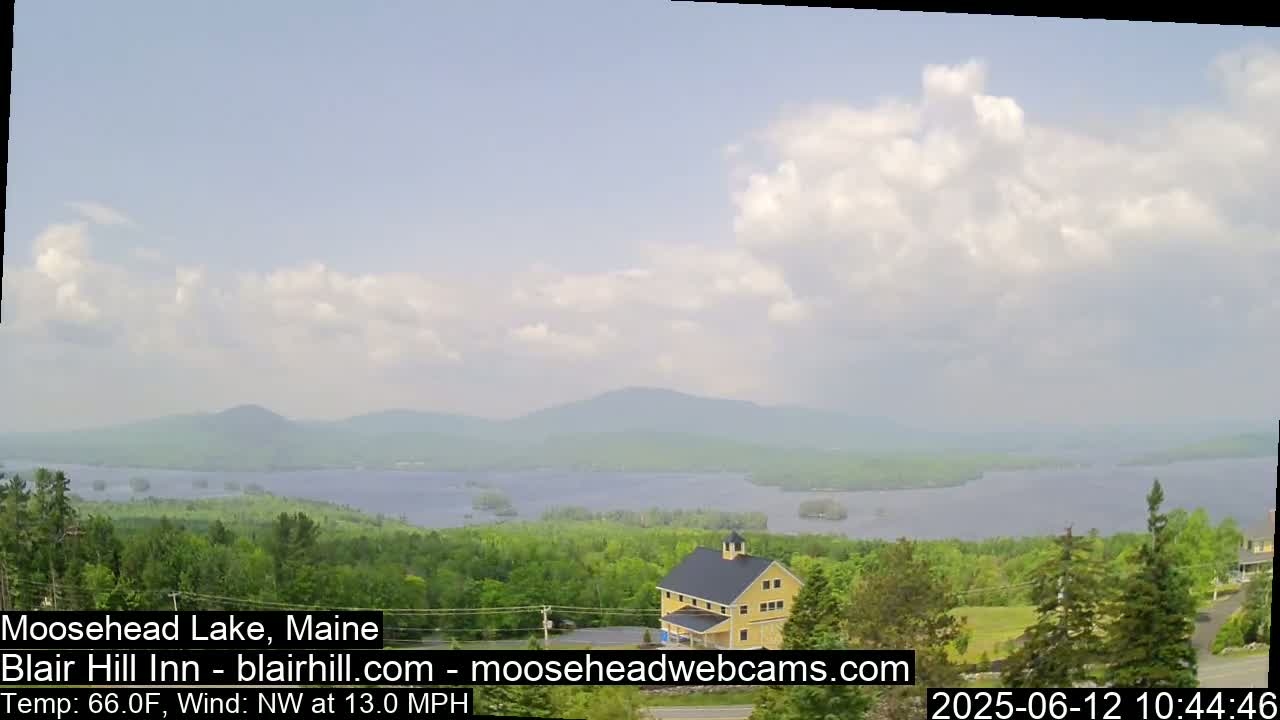 A partly cloudy sky overlooks a large lake, surrounded by green trees and distant mountains, with a yellow building visible in the foreground.