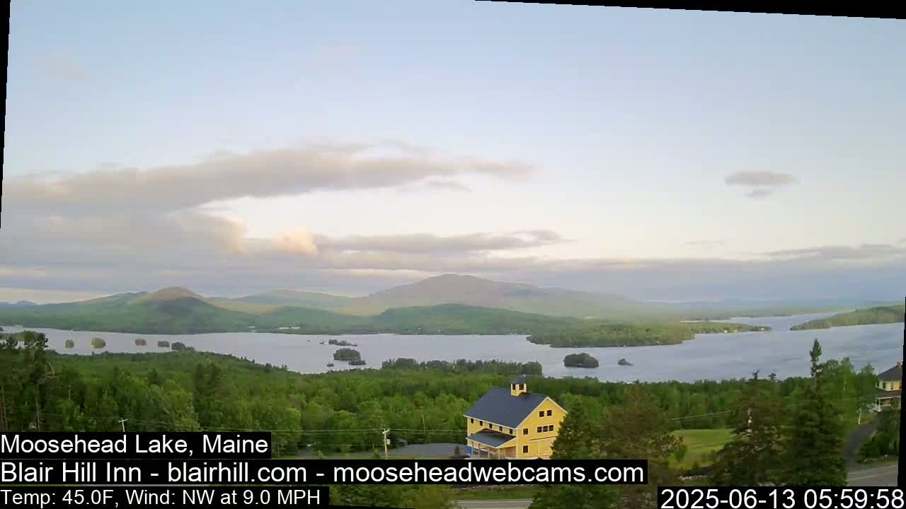 A calm, partly cloudy morning reveals a tranquil lake nestled among verdant hills and mountains, with a yellow building visible in the foreground.