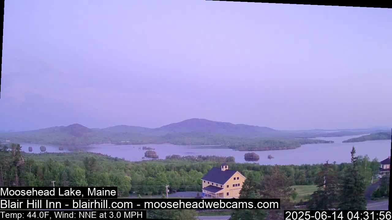 A calm, twilight view of Moosehead Lake, featuring a yellow building nestled among green trees and a distant mountain range under a pale purple sky.