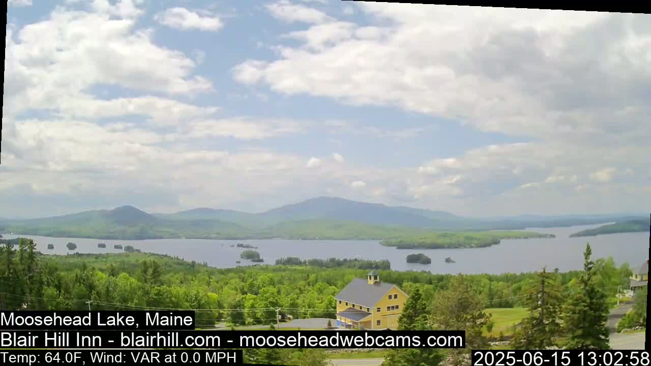 A partly cloudy day reveals a view of a lake surrounded by green trees and distant mountains, with a yellow barn visible in the foreground.