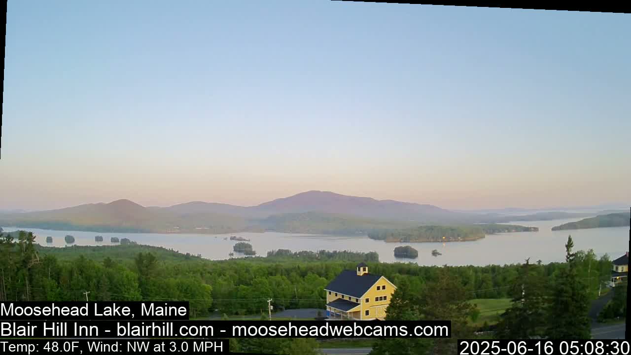 A calm, sunrise scene shows a large lake surrounded by green trees and mountains under a pale blue sky.