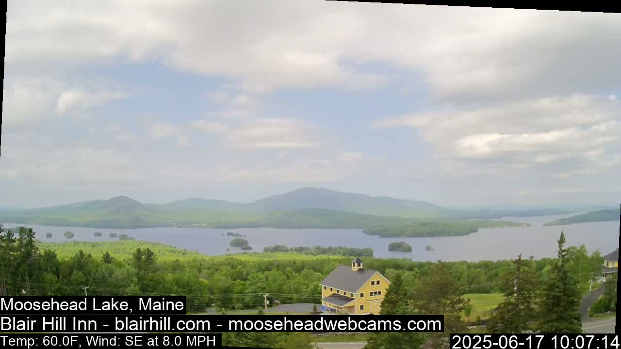 A yellow building sits on a hill overlooking a large lake with islands and distant mountains under a partly cloudy sky.