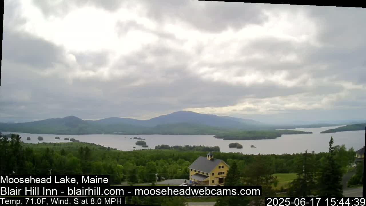 A large lake is seen from an elevated vantage point under a mostly cloudy sky, with a yellow building and green trees in the foreground and mountains in the distance.