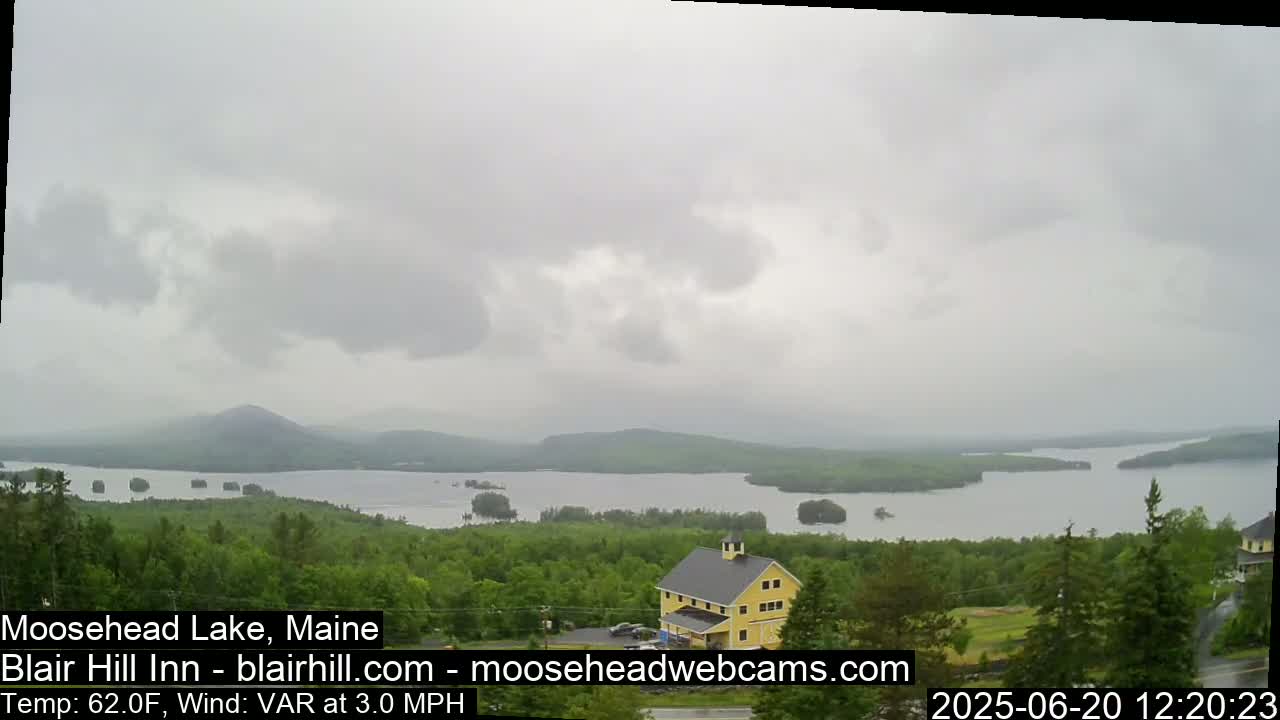A yellow building sits on a hill overlooking a large lake surrounded by green trees under an overcast sky.