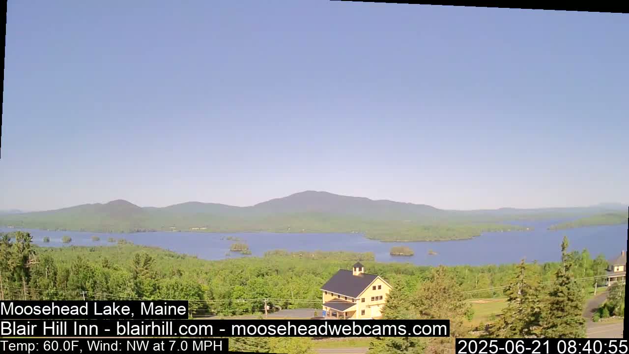 A large lake with a distant mountain range under a clear blue sky is viewed from a high vantage point, showing a building and green trees in the foreground.