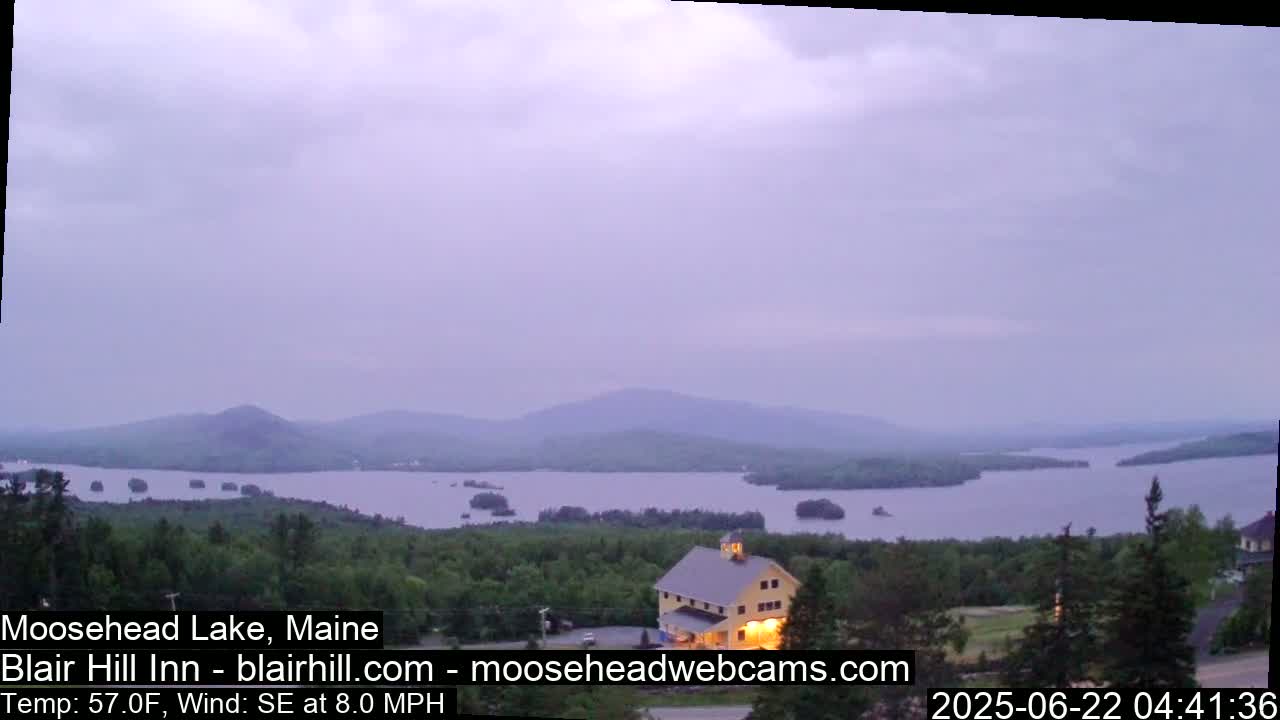 A yellow barn sits atop a hill overlooking a calm lake and distant mountains under a hazy, overcast sky.