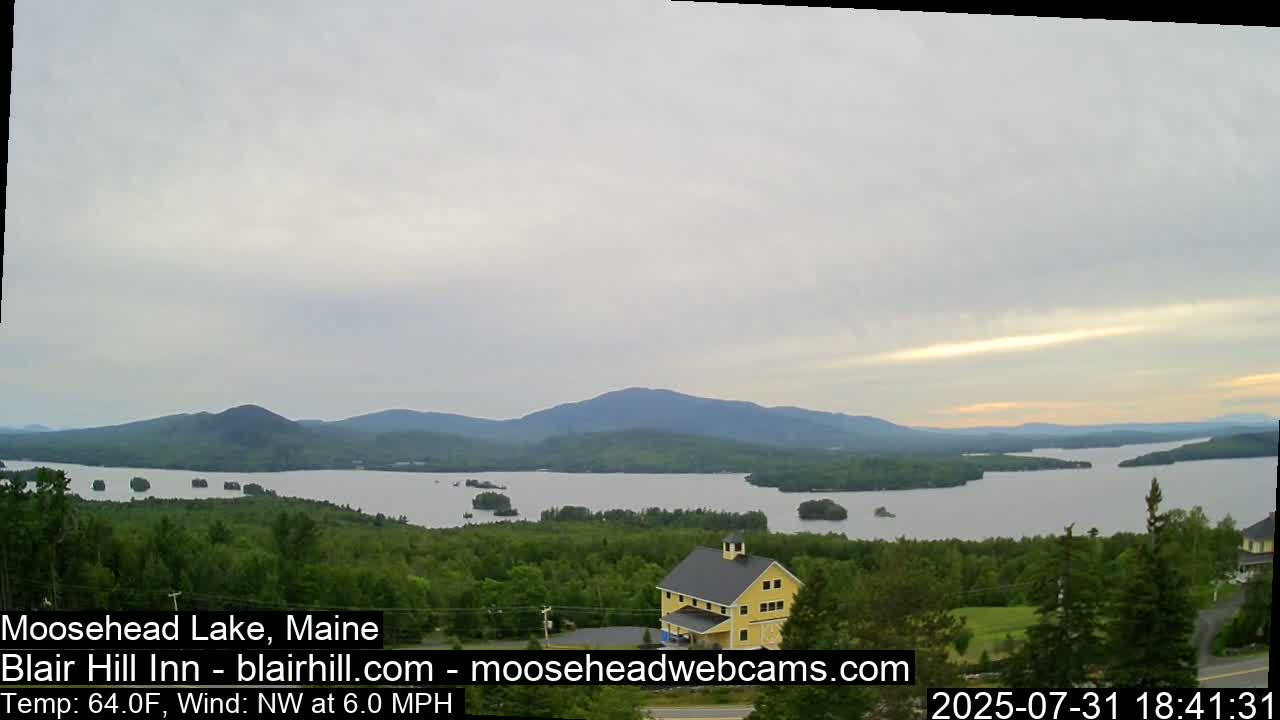 A large lake surrounded by green hills and a yellow building is visible under an overcast sky with a light northwest wind.