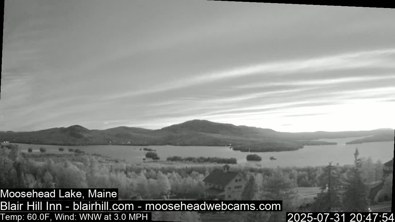 A grayscale view of a calm lake at sunset, nestled between gently rolling hills under a partly cloudy sky.