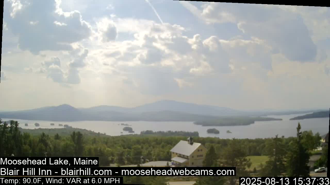 A partly cloudy day with a 6 mph variable wind shows a view of a large lake with islands and distant mountains,  from a vantage point overlooking a building nestled among green trees.