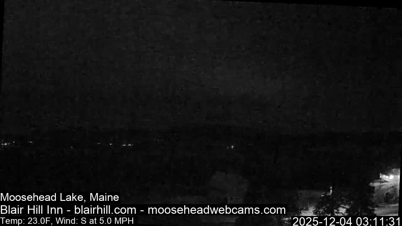 A predominantly dark and blurry nighttime image shows a distant horizontal line of faint lights, possibly across a body of water or valley, with some extremely dim shapes and a brighter light source in the foreground, and no other discernible weather conditions.