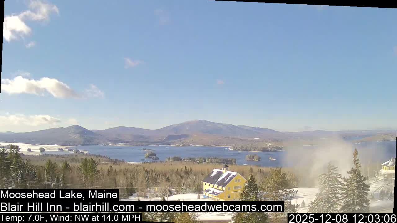 A clear and sunny winter day illuminates a snow-covered landscape featuring a wide lake dotted with islands, framed by distant mountains and dense evergreen forests, with a prominent yellow building and a cloud of blown snow visible in the foreground.