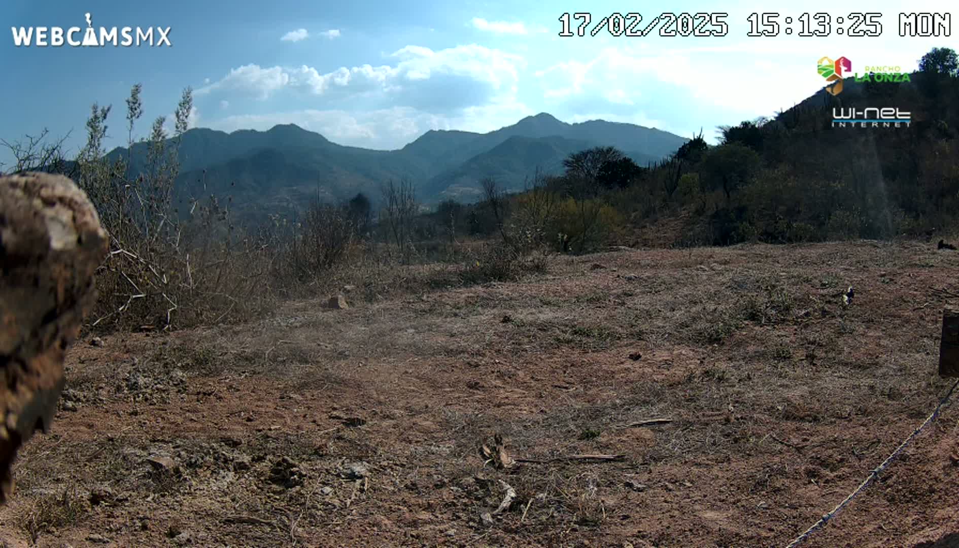 A mostly barren, dry, reddish-brown dirt field extends to a background of distant mountains under a partly cloudy sky.