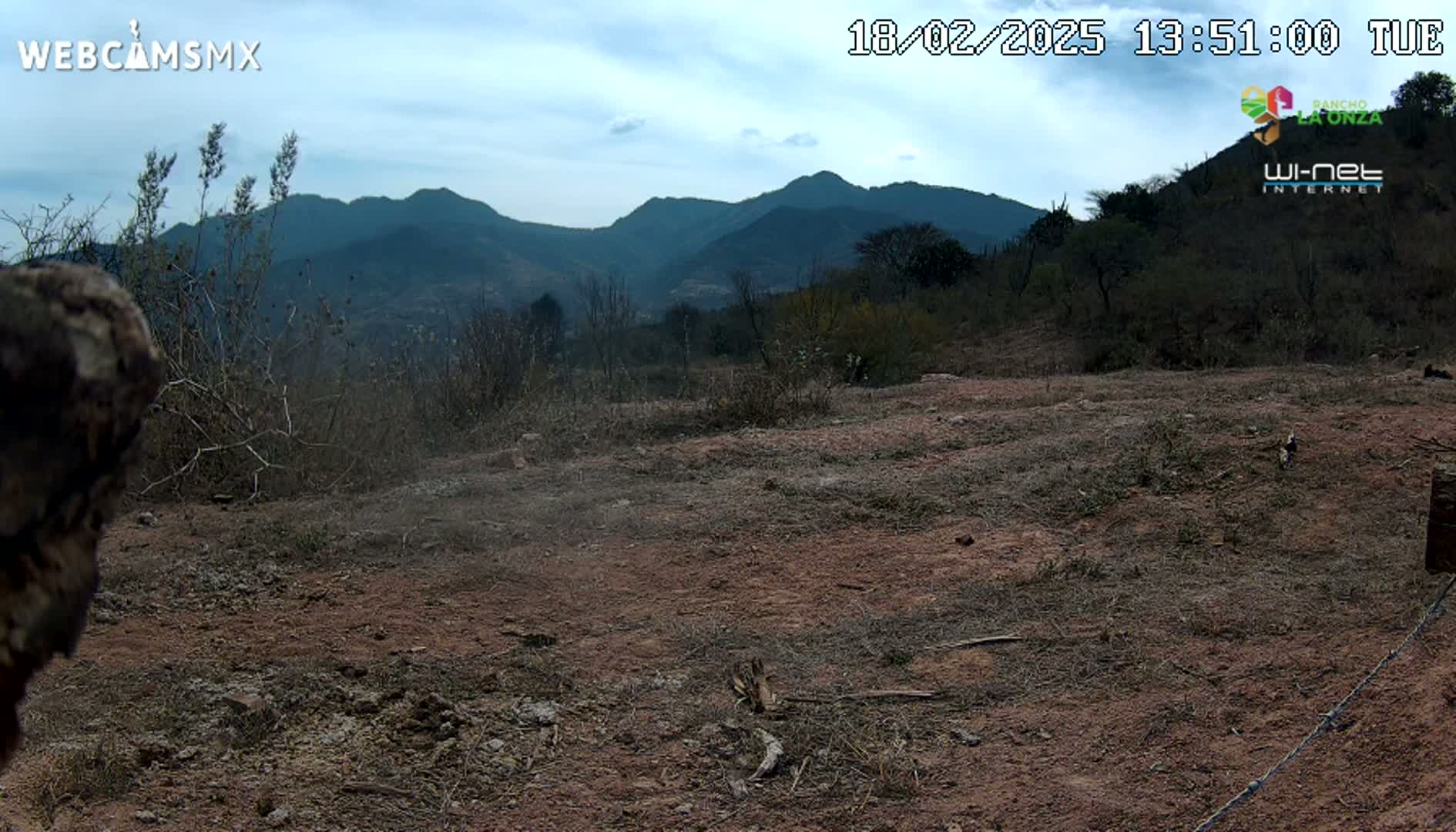 A mostly barren, reddish-brown dirt field stretches toward a range of dark mountains under a mostly clear, light-blue sky.
