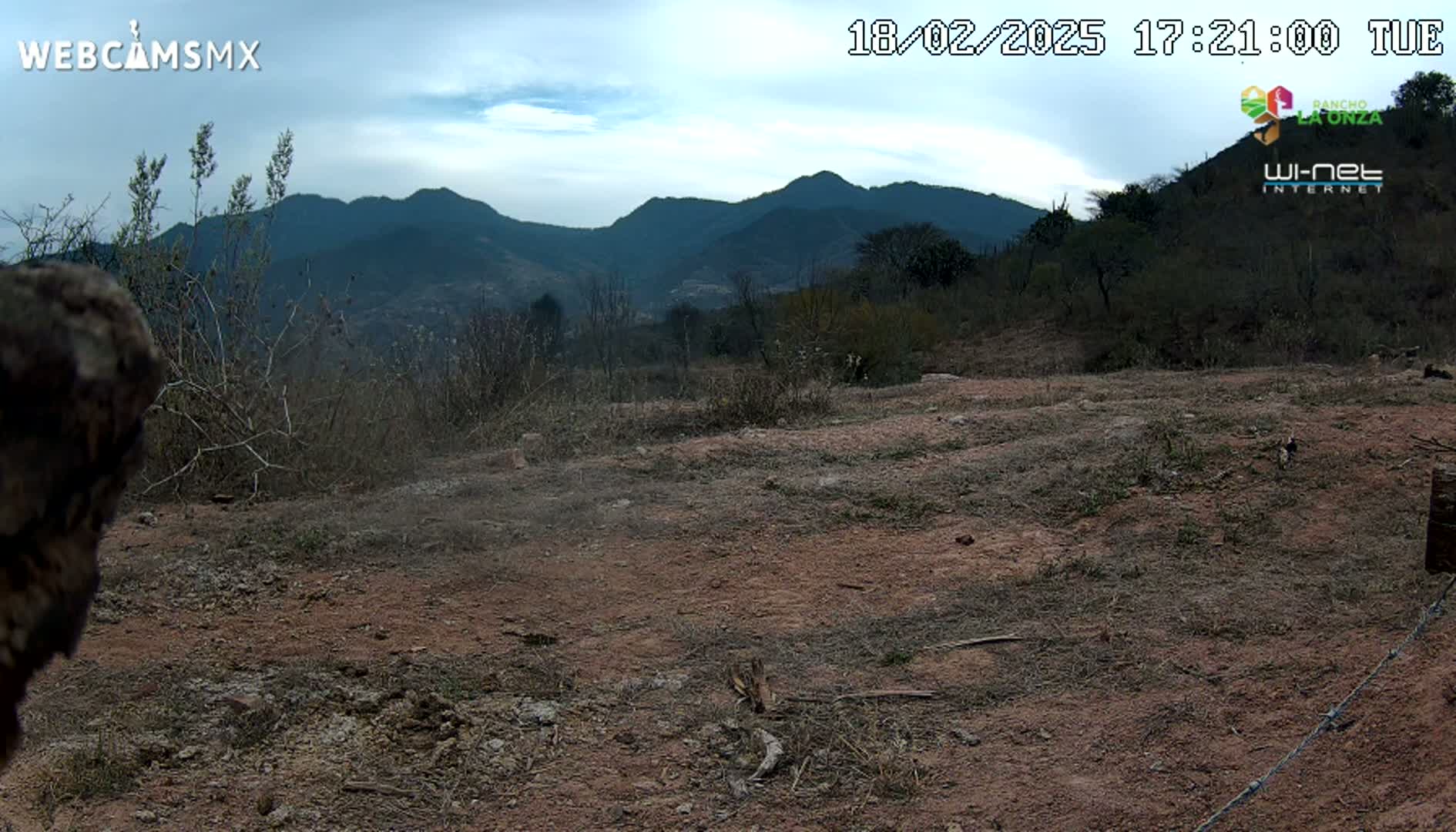 A mostly barren, dry, reddish-brown dirt field stretches out before a range of dark, distant mountains under a partly cloudy sky.