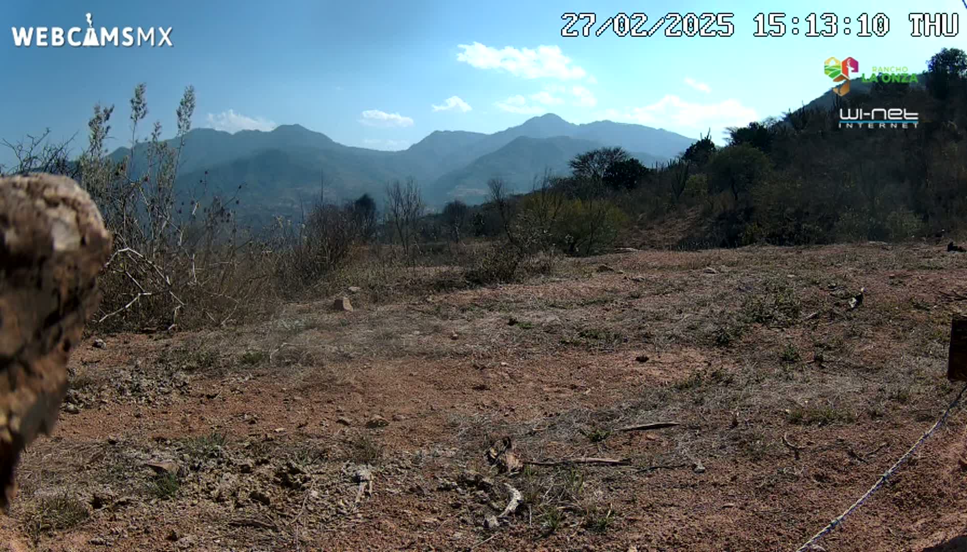 A mostly barren, reddish-brown dirt field stretches towards a background of distant mountains under a partly cloudy, sunny sky.