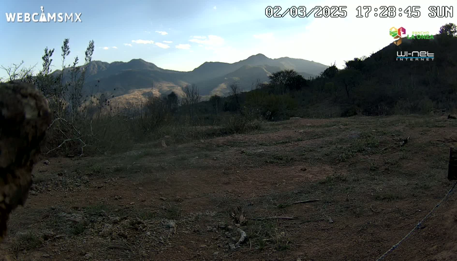 A mostly barren, dirt landscape with sparse brush and distant mountains under a partly cloudy sky.