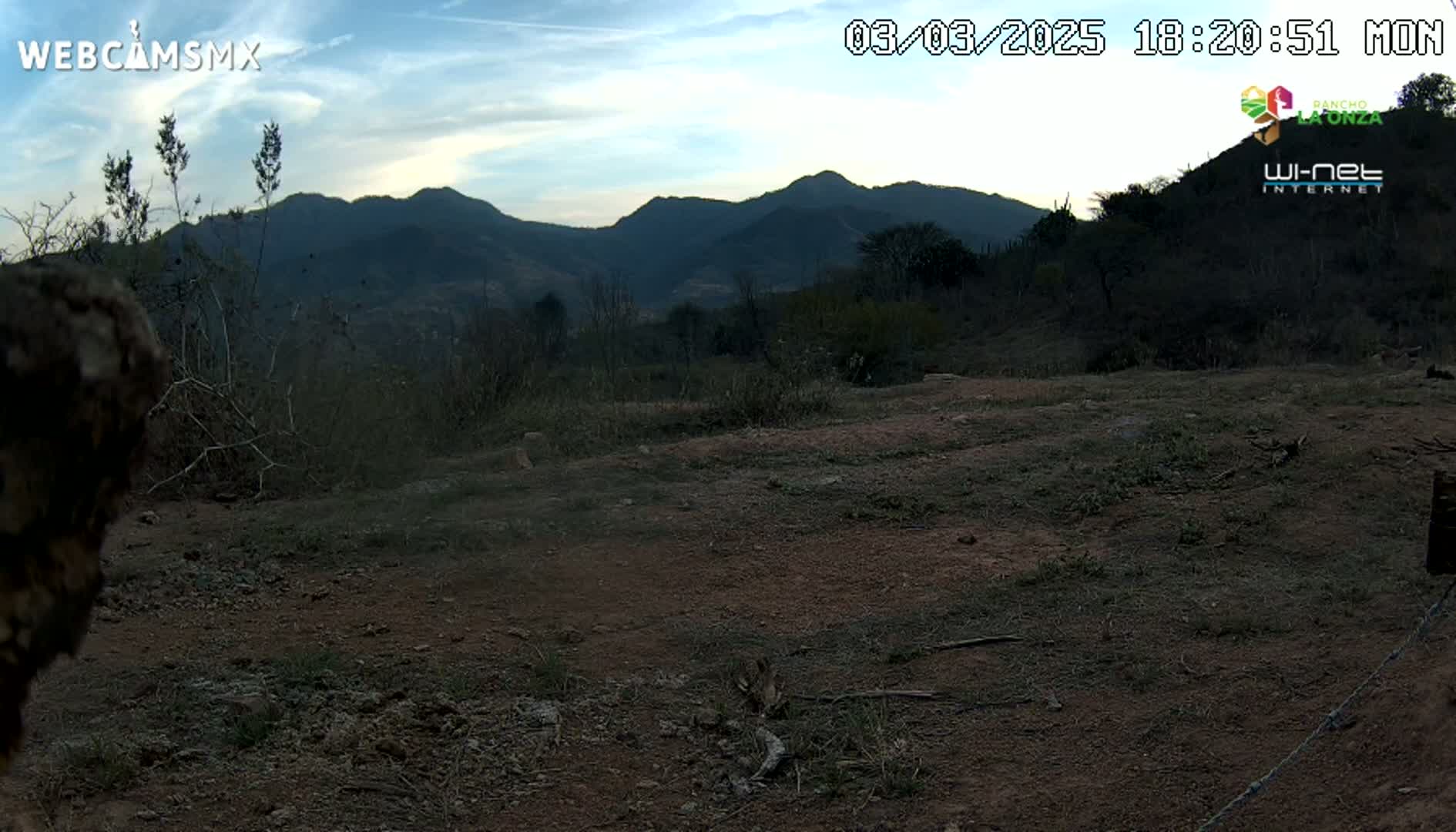 A dirt field with sparse vegetation and a barbed wire fence is in the foreground, with mountains in the background under a partly cloudy twilight sky.