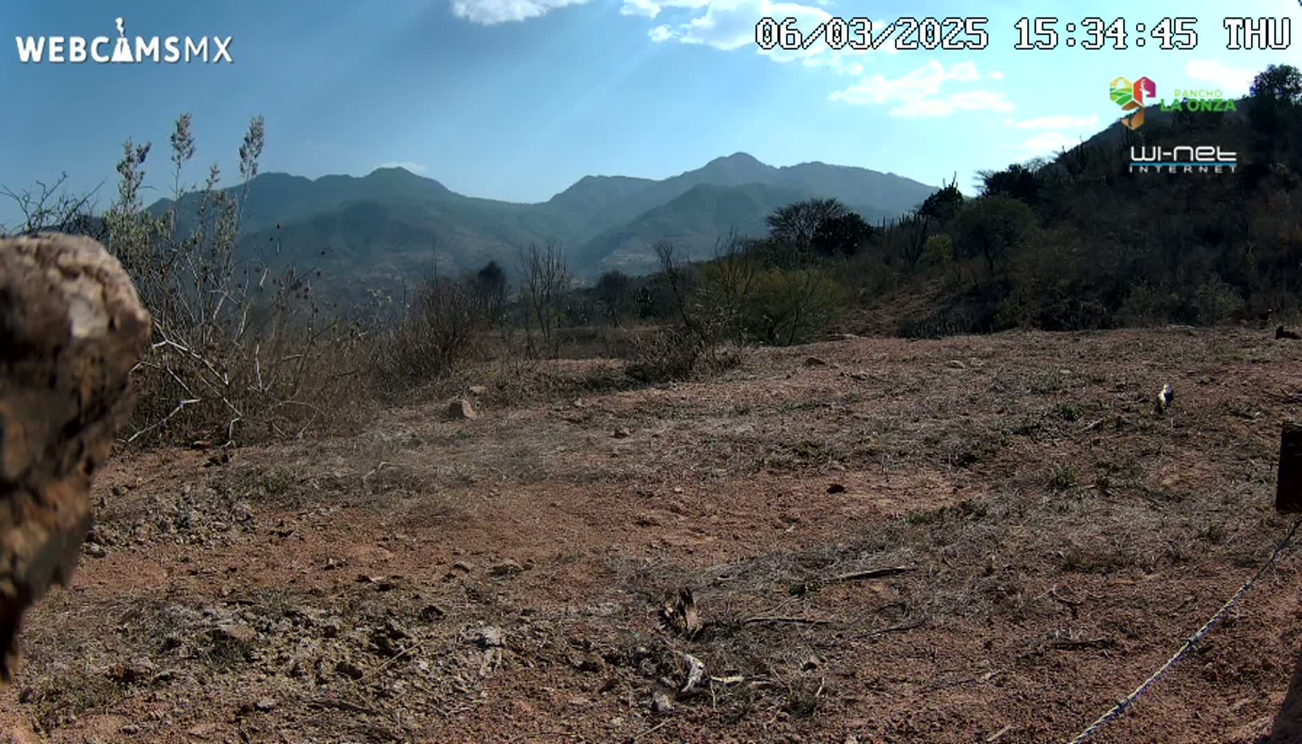 A mostly barren, reddish-brown landscape with sparse, dry brush and distant mountains under a mostly clear, sunny sky.