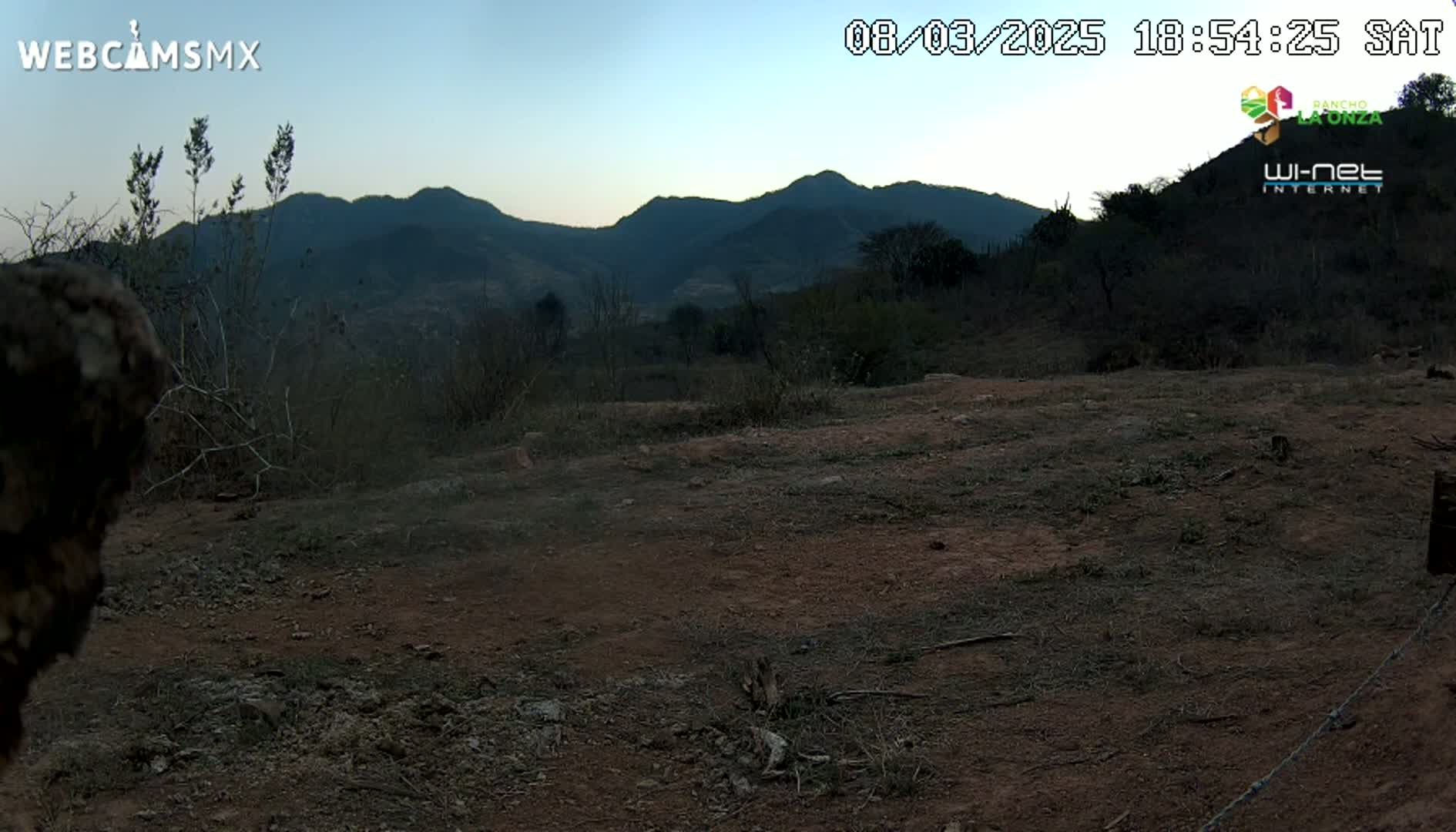A flat, barren expanse of reddish-brown dirt and sparse, dry brush stretches towards a range of dark, distant mountains under a twilight sky.