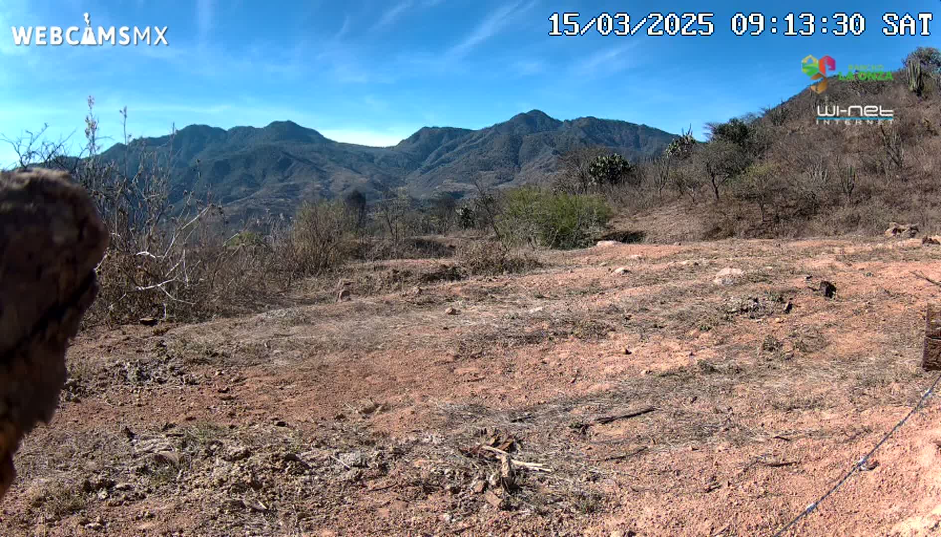 A mostly barren, reddish-brown dirt landscape stretches to a range of mountains under a clear blue sky.