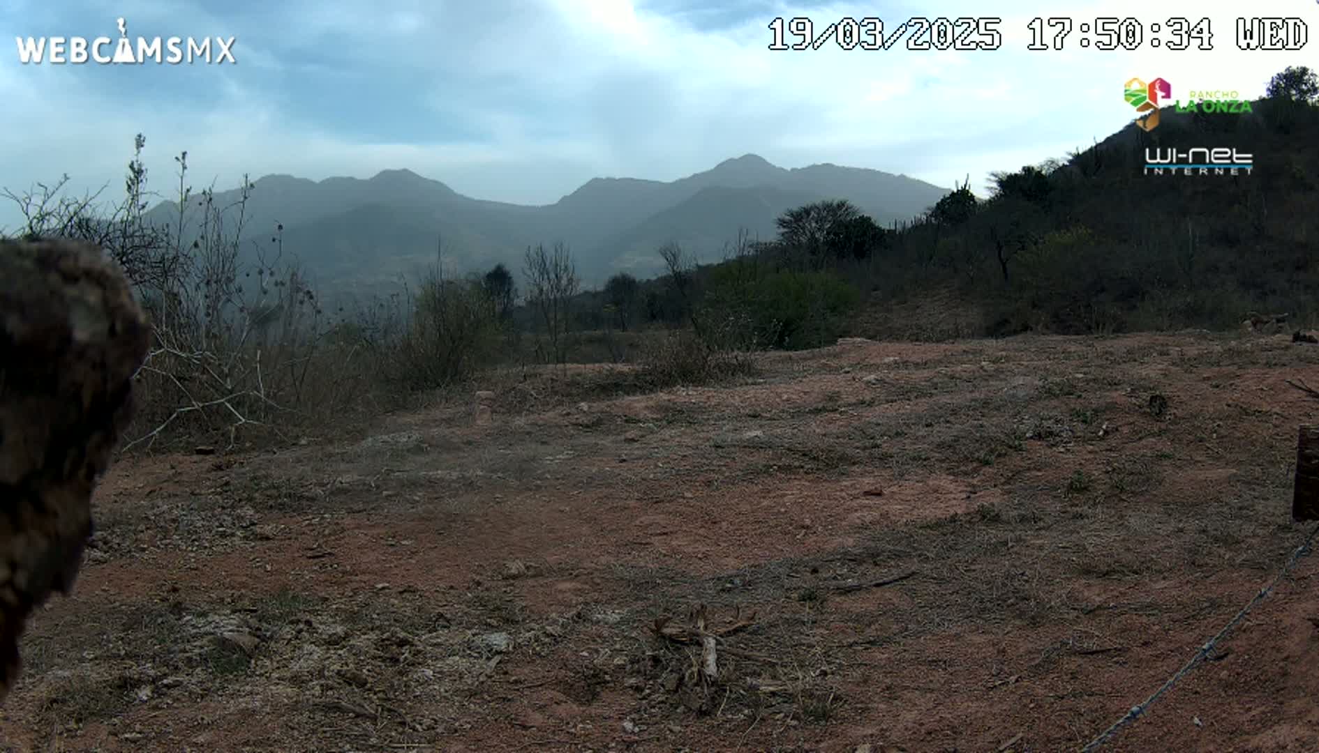A mostly barren, reddish-brown dirt expanse stretches out before a sparsely vegetated hillside under a partly cloudy sky.