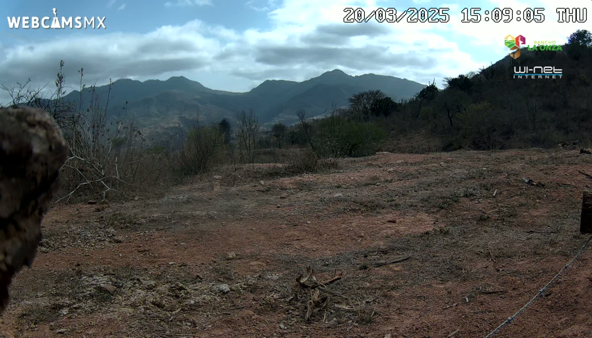 A partly cloudy day reveals a dry, reddish-brown expanse of land leading to a range of dark, distant mountains.