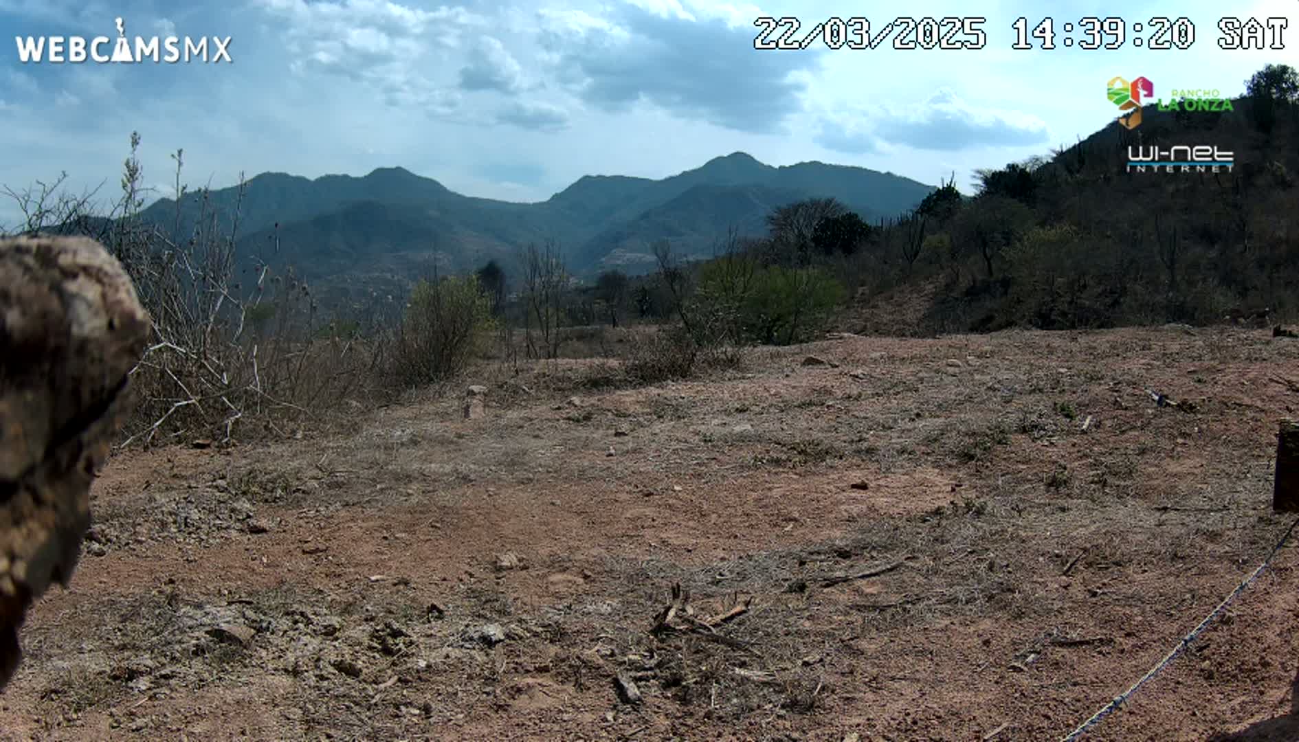 A partly cloudy day reveals a dry, reddish-brown landscape with sparse vegetation in the foreground, leading to a vista of distant, dark-blue mountains.
