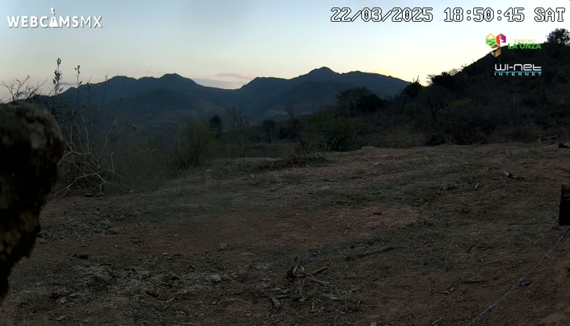 A twilight view shows a dry, dirt ground in the foreground, sparsely vegetated hills in the mid-ground, and mountains silhouetted against a pale dusk sky in the background.