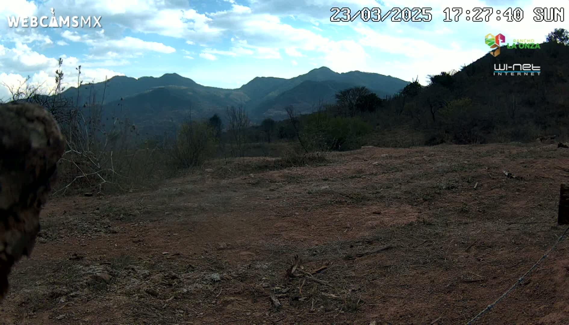 A partly cloudy day reveals a view of brown, barren land in the foreground, with scrubby vegetation and dark mountains in the background.